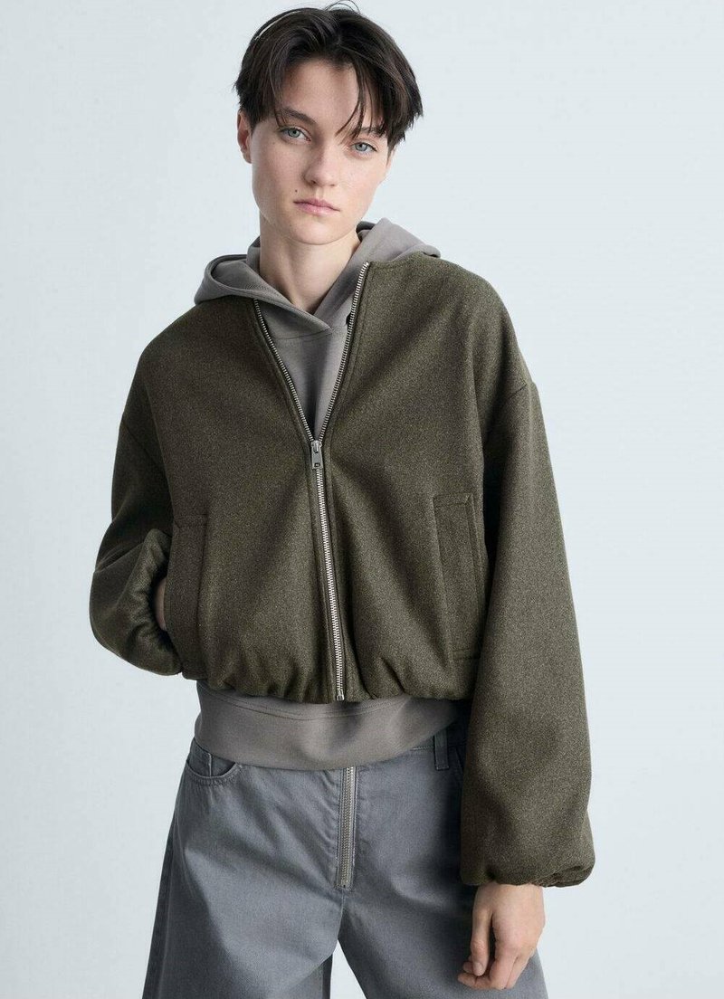 Giacca bomber verde oliva con zipper frontale, spalle scese e orlo a costine. Indossata sopra un hoodie grigio, abbinata a pantaloni grigi.