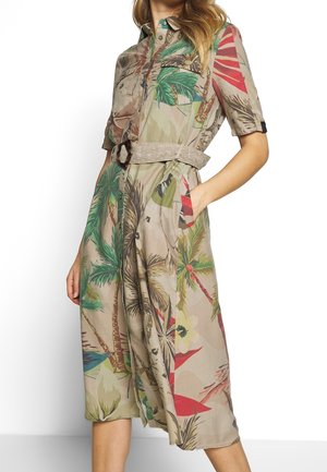 Frau trägt ein tailliertes, beiges Hemdkleid mit grünen und roten tropischen Blättern und Palme-Drucken, Hand in der Tasche.