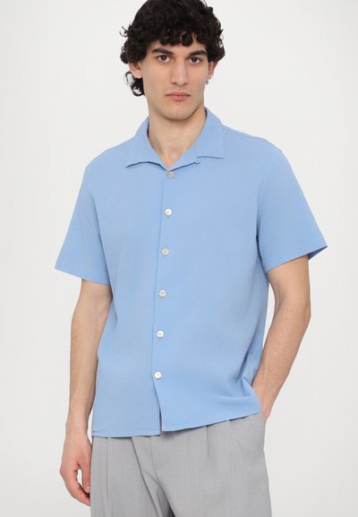 Camicia a maniche corte in tessuto azzurro chiaro con una texture liscia, caratterizzata da un colletto e sei bottoni bianchi sul davanti.