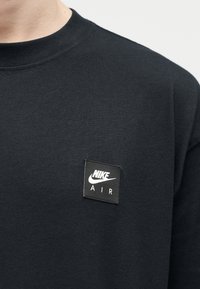 Μαύρο t-shirt Nike Air με μικρό τετράγωνο λογότυπο στο πάνω μέρος του στήθους, που δείχνει το σήμα Nike Swoosh και το κείμενο "AIR".