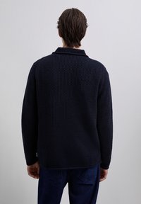 Homme vu de dos portant un pull long à manches longues en navy texturé et un pantalon bleu foncé, sur un fond gris clair uni.