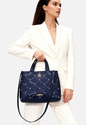 Bolso acolchado de color azul marino con adornos dorados y un logo, que cuenta con dos asas y una correa de hombro desmontable. Fabricado con material brillante.