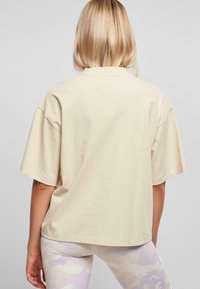 Světlý béžový oversized tričko vyrobené z měkké tkaniny s volným střihem a krátkými rukávy, s límečkem vzadu a hladkým designem.