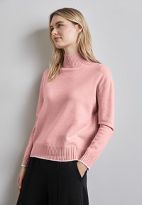 Rosa Rollkragenpullover mit geripptem Kragen und Saum, weiche Textur, lässige Passform, kombiniert mit schwarzen Hosen, Modell posiert entspannt mit Händen in den Taschen.