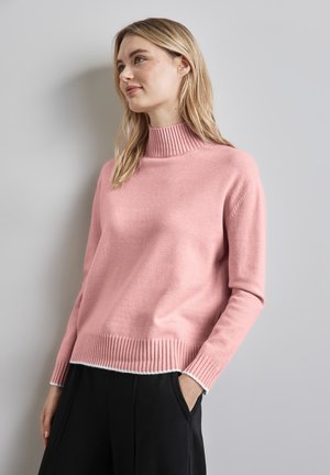 Różowy sweter z golfem, z prążkowanym kołnierzem i dołem, miękka faktura, luźny krój, w połączeniu z czarnymi spodniami, modelka swobodnie pozuje z rękami w kieszeniach.