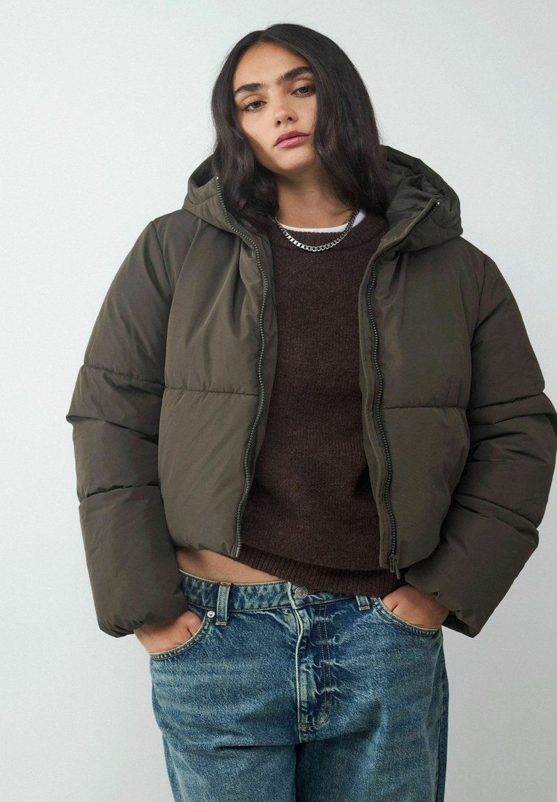 Olivgrüne Daunenjacke mit Kapuze, Reißverschluss und elastischen Bündchen, über einem braunen Pullover getragen und kombiniert mit blauen Jeans.