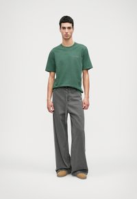 Zaļš īsām piedurknēm t-krekls ar logo, apvienots ar platām pelēkām džinsām. Iekļautas brūnas kurpes, stāvot uz gaiša fona.