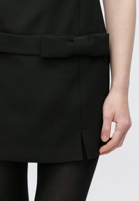 Vestido preto com um laço na cintura, feito de tecido texturizado. Detalhe de fenda lateral visível, usado com collants pretos.