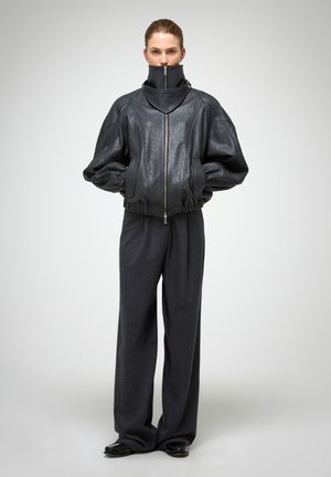 Black leather bomber jacket with a high collar, zip closure, and gathered cuffs, paired with wide-leg dark gray trousers.