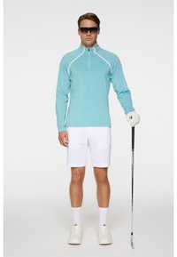 Ljusblå långärmad polo med halvdragkedja och vita detaljer, i kombination med vita shorts. Modellen håller en golfklubba och har på sig solglasögon och handskar.