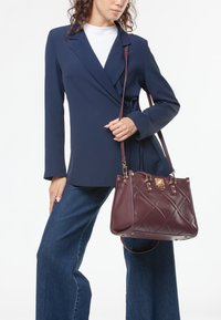 Bolso acolchado marrón con herrajes dorados, llevado al hombro. Modelo con blazer azul marino y camisa blanca, combinado con jeans azules acampanados.