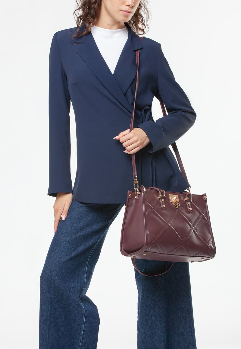 Bolso acolchado marrón con herrajes dorados, llevado al hombro. Modelo con blazer azul marino y camisa blanca, combinado con jeans azules acampanados.