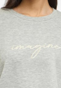 Grå t-skjorte med korte ermer laget av mykt, teksturert stoff, med brodert tekst "imagine" i kremfarge over brystet.