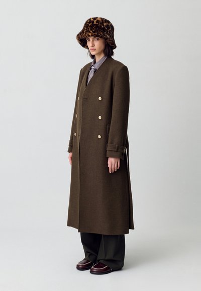 Cappotto in lana verde oliva a doppio petto con bottoni dorati, tasche laterali e taglio lungo. Abbinato a un cappello a secchiello con stampa leopardo marrone.