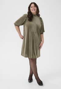 Olijfgroene geribbelde jurk met hoge hals, korte pofmouwen en een relaxte pasvorm. Glanzende zwarte puntige schoenen en doorschijnende maillots.