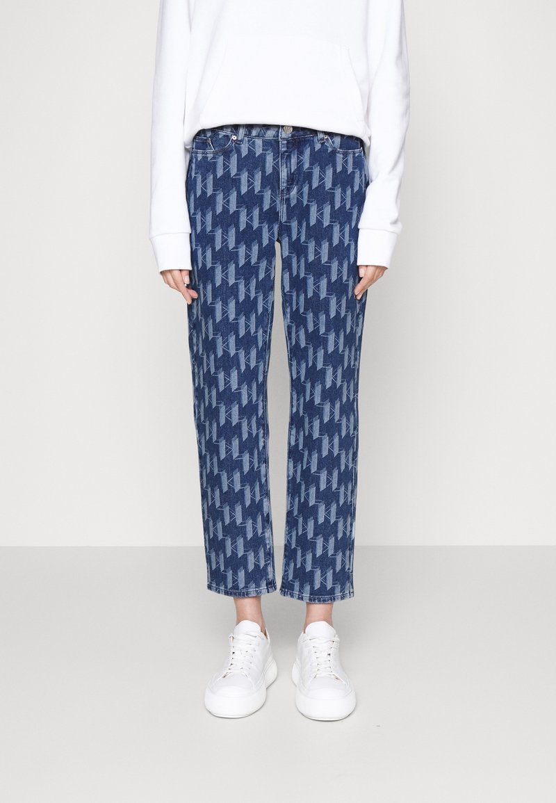Personne portant un jean droit bleu à motif, un sweat-shirt blanc et des baskets blanches, debout devant un fond clair uni.