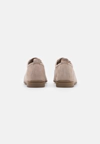 Scarpe in camoscio di colore beige chiaro con retro arrotondato, caratterizzate da una superficie liscia e una suola in gomma marrone. Nessun lacci o accessori visibili.
