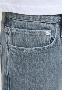 Los jeans de mezclilla azul claro presentan un primer plano de un bolsillo con botones metálicos y costuras visibles, que muestran una superficie de tela texturizada.