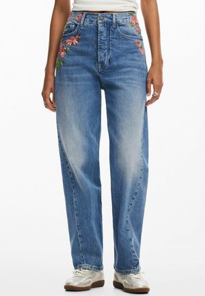 Hochtaillierte blaue Jeans mit roten und grünen Blumenstickereien auf den Taschen, kombiniert mit weißen Sneakers und einem in die Hose gesteckten weißen Oberteil.