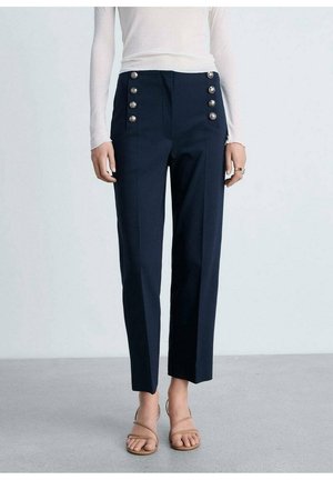 Marineblaue, hochtaillierte Hose mit sechs silbernen Knöpfen, weißes Langarmoberteil und beige Sandalen mit Riemchen an einer stehenden Person.