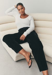 Sorte kordfløjlsbukser med en afslappet pasform, kombineret med en hvid langærmet top, på en tekstureret lysfarvet sofa. Sorte sandaler med stropper.