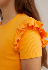 Dzeltena ribota kokvilnas T-krekls ar volāniem īsajās piedurknēs; gluda tekstūra ar pieguļošu piegriezumu.