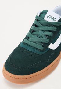 Sapatilhas de camurça verdes com o logótipo Vans em branco, atacadores texturizados, perfurações na ponta e sola de borracha castanho-claro.