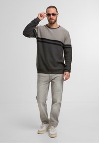 Grau und schwarz gestreifter Strickpullover mit Rundhalsausschnitt, kombiniert mit hellgrauen Jeans und weißen Turnschuhen. Das Modell trägt Sonnenbrillen.