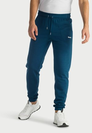 Man wearing blue Bench sweatpants with drawstring waist and white sneakers, standing against a plain light background.