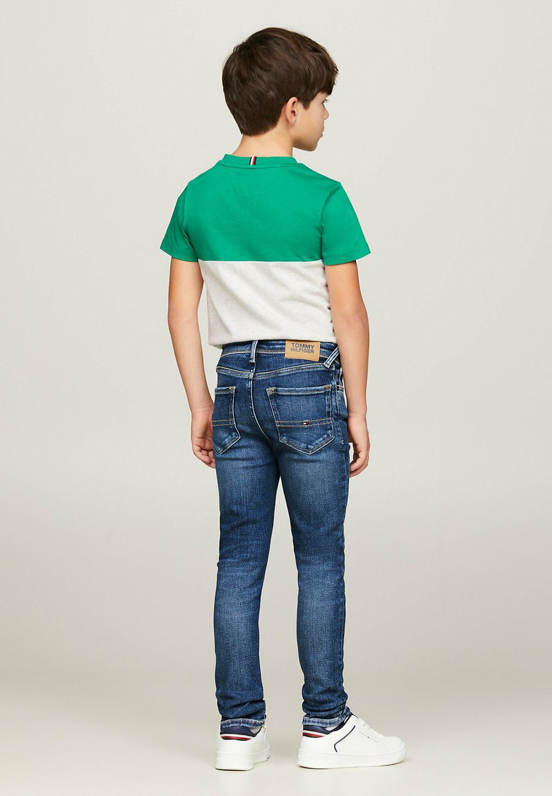 Zielono-szaro zblokowana koszulka, w połączeniu z ciemnoniebieskimi skinny jeansami, z naszywką z logo na plecach. Białe sneakersy z kolorowymi akcentami.