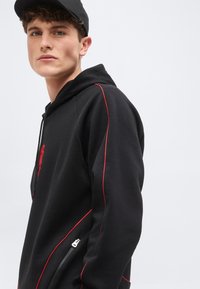 Νεαρός άνδρας που φοράει μαύρο φούτερ με κόκκινες ραφές και μαύρο καπέλο, κοιτάζοντας ελαφρώς πλαγίως με φόντο μια απλή ανοιχτόχρωμη επιφάνεια.