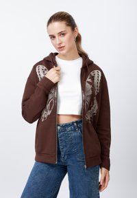 Brūns rāvējslēdzēja džemperis ar tauriņa grafiku, izgatavots no kokvilnas maisījuma auduma ar ribas aprocēm. Valkāts kopā ar īsu baltu topu un zilām džinsām.