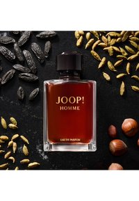 Klar glassflaske med Joop! Homme-parfyme med en mørk væske, svart kork og gulltekst. Omgitt av krydder: kardemomme, tonkabønner og hasselnøtter.