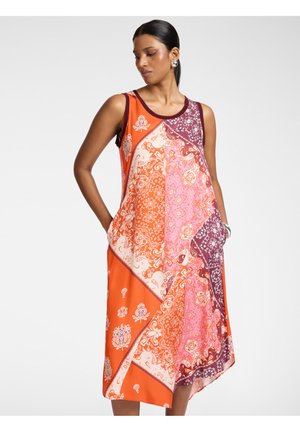 Kvinde med mørkt hår i hestehale iført en ærmeløs patchwork-kjole med orange, pink og mørkerøde blomsterprint, hænderne i lommerne, på hvid baggrund.