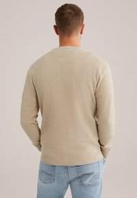 Beige textúrájú pulóver kerek nyakkal és hosszú ujjakkal, bordás aljjal, világoskék farmernadrággal párosítva.