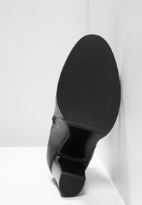 Botte en cuir noir à la cheville inclinée contre un coin blanc, semelle tournée vers la caméra, montrant un talon texturé et une semelle lisse.