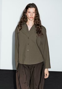 Olive green button-up shirt with a collar, long sleeves, and relaxed fit, paired with dark brown pleated trousers. Smooth cotton fabric.