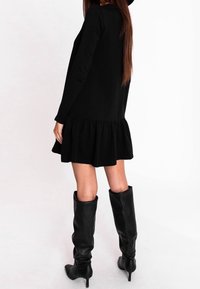 Czarna sukienka z długim rękawem, z falbaną u dołu, wykonana z miękkiego materiału. Modelka zestawia ją z wysokimi czarnymi skórzanymi botkami na obcasie.