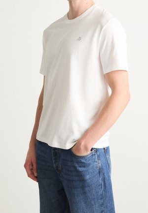 Homme portant un t-shirt blanc à manches courtes avec un petit logo « M OP » et un jean bleu, main dans la poche, sur un fond clair uni.