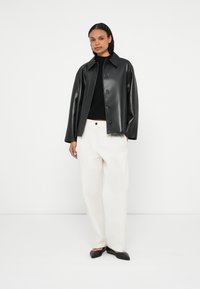 Sort jakke af kunstlæder med krave og knaplukning, kombineret med hvide bukser med vide ben og sorte spidse flade sko.