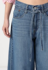 Pantalones de mezclilla azul con un diseño de pernera ancha, que cuentan con un detalle de lazo en la parte delantera, trabas para cinturón y el clásico estilo de cinco bolsillos. Textura suave y acabado desgastado.