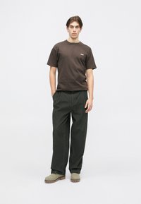 Brun bomulds T-shirt med et lille hvidt logo, parret med mørkegrønne fløjlbukser og beige slip-on sko. Enkel, afslappet påklædning.