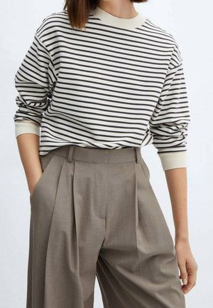 Kvinde iført beige plisserede bukser og en cremefarvet langærmet sweater med sorte vandrette striber, hænderne i lommerne.
