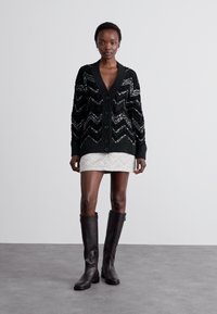 Μαύρο καρκινάκι με ζιγκζαγκ μοτίβο και παγιέτες, διαθέτει V-λαιμόκοψη, κουμπί κλείσιμο και ριμπ μανσέτες. Συνδυασμένο με λευκή υφασμάτινη μίνι φούστα και μπότες μέχρι το γόνατο.