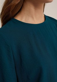 Blågrön blus med rund halsringning, tillverkad av lättviktigt material, med en texturerad yta och avslappnad passform.