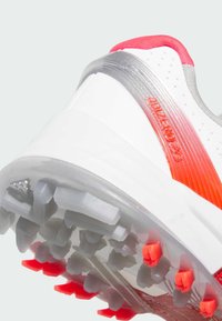 Witte sportschoen met rode en zilveren accenten, met een gestructureerde rubberen buitenzool voorzien van kegelvormige spijkers voor optimale grip.
