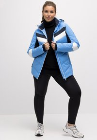 Ljusblå vadderad jacka med svart och vitt zigzagmönster, huva, dragkedja framtill och elastiska ärmslut, i kombination med svarta leggings och sneakers.