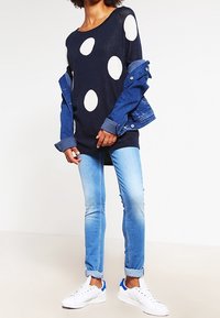 Pull marine avec de grands pois blancs, superposé à une veste en jean. Jeans bleu clair et baskets blanches avec des accents bleus.