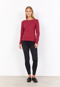 Red and burgundy striped long-sleeve top, round neckline, paired with black skinny jeans and black loafers, standing on a neutral floor.