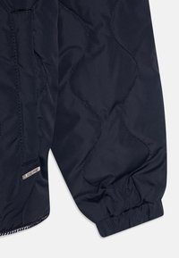 Secção de casaco puffer azul marinho escuro com padrão acolchoado, punhos elásticos e um pequeno emblema metálico da marca no tecido.